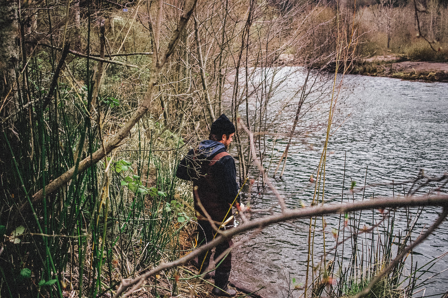 Person fishing with a backpack walking along a path by a river in a forested area